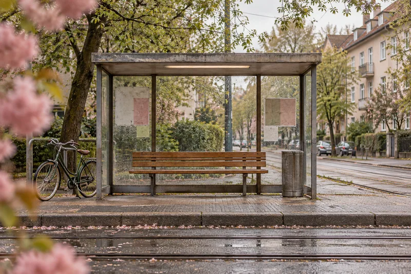 Eine schlichte Haltestelle mit Bank und Glasdach an einer baumbestandenen Wohnstraße.